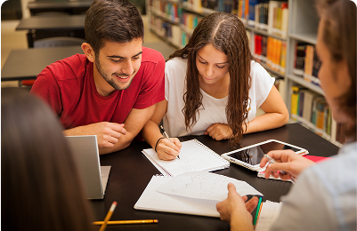 Students sitting together in a library, discussing and studying with notebooks and laptops.